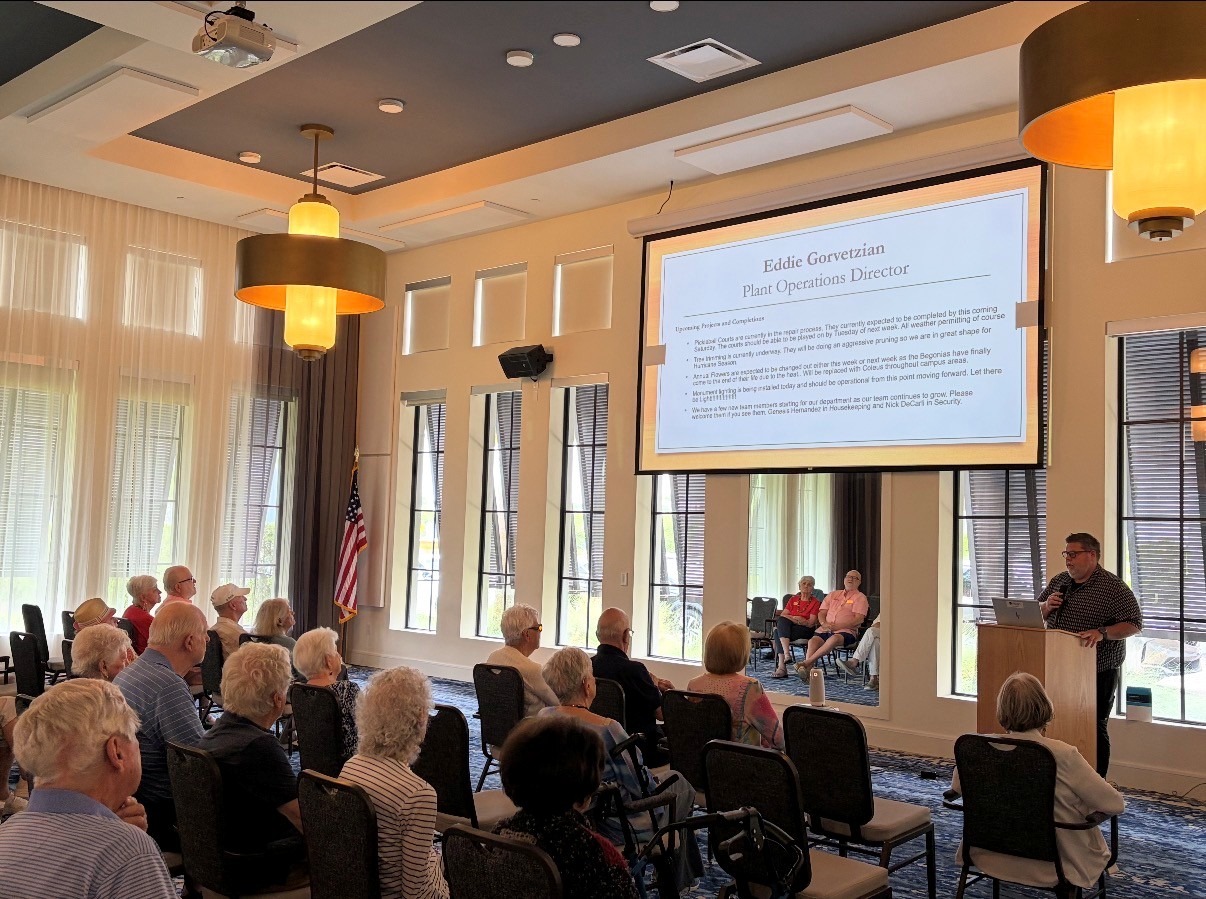 Wellington Bay Plant Operations Director presenting a hurricane readiness slide to a seated audience of senior residents inside a brightly lit luxury community clubhouse.