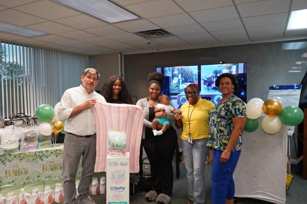 (Left to right) Owen O'Neill, Lottie Moise, MSW, a mother with her baby, Akeelah Jackson, and Angela Kelly celebrate providing safe sleeping arrangements for infants at the Bundles of Joy event. Owen O'Neill, Lottie Moise, Akeelah Jackson holding a baby, and Angela Kelly posing together indoors by a Cribette box and baby supplies.