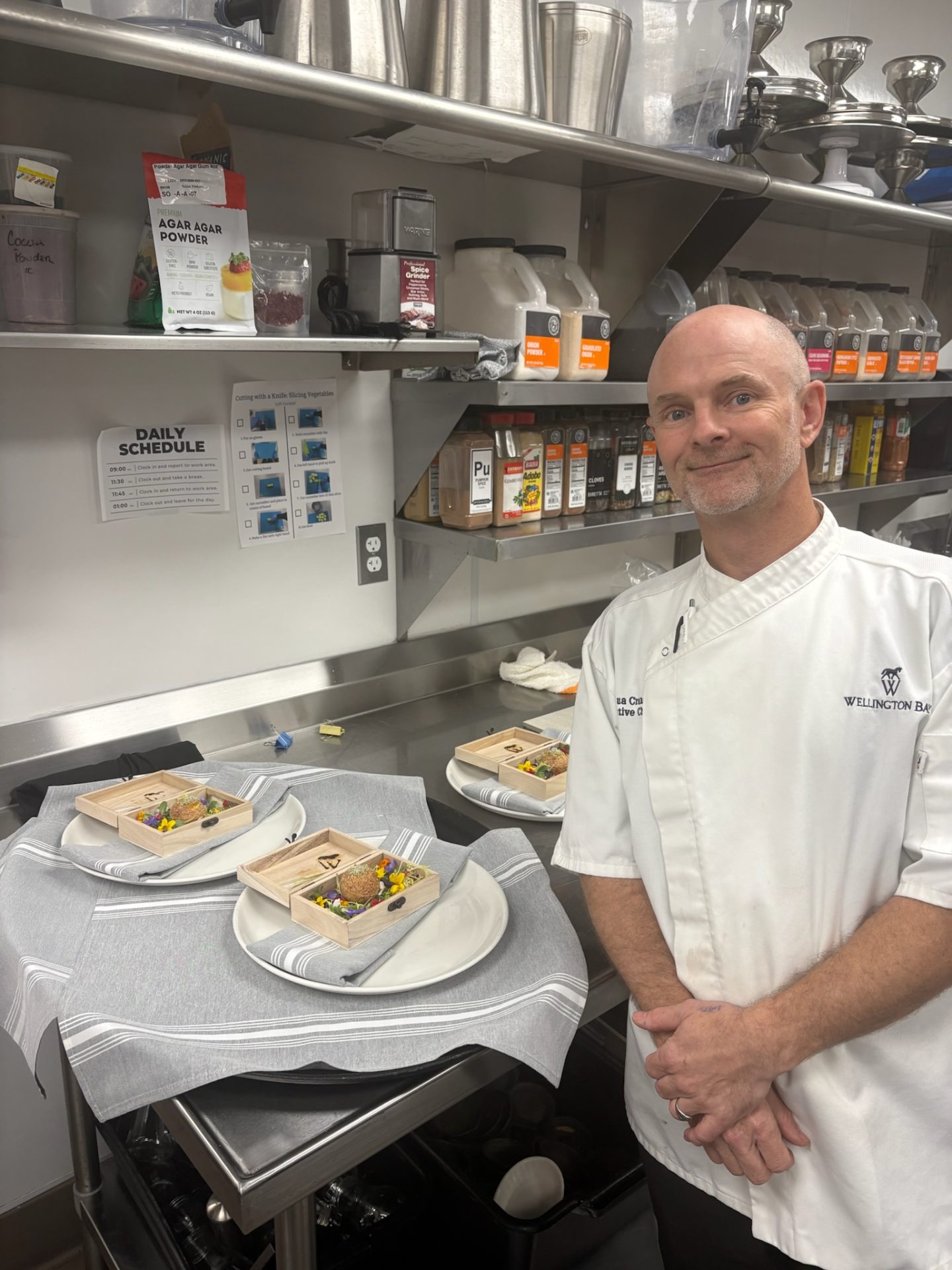 Chef Joshua Crane of Wellington Bay standing proudly in a commercial kitchen next to plated portions of his gourmet Pani Puri entry.
