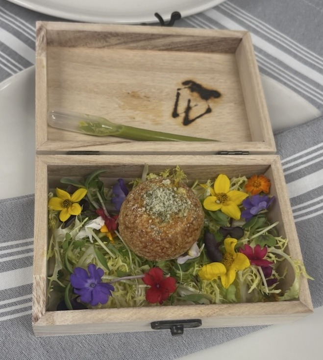 A single gourmet Pani Puri resting on a bed of microgreens and colorful edible flowers inside a small, open wooden box with a cilantro-honey dressing pipette.