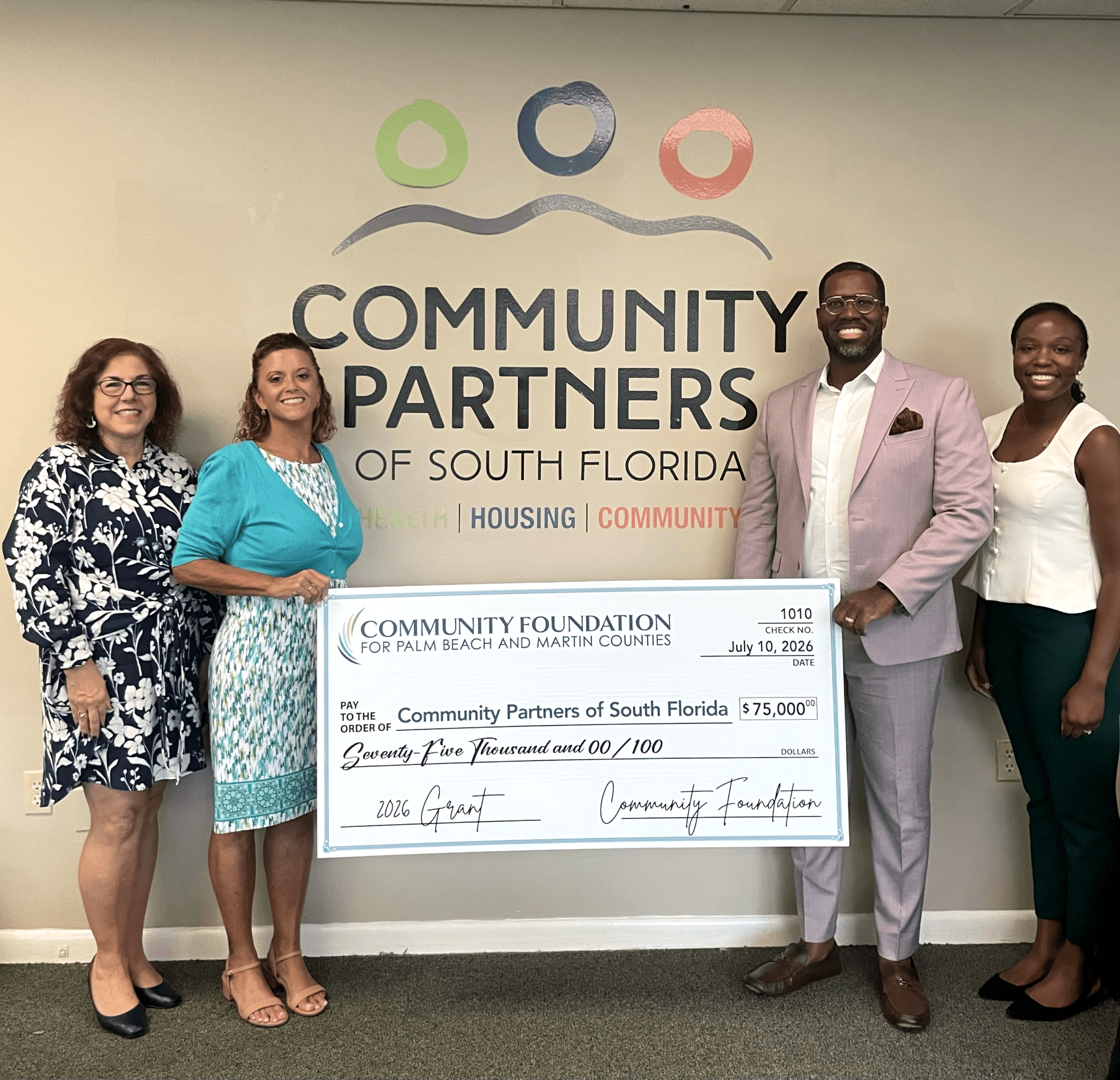 Four professionals standing in front of a Community Partners of South Florida wall logo, holding a large $75,000 ceremonial grant check dated July 10, 2026.