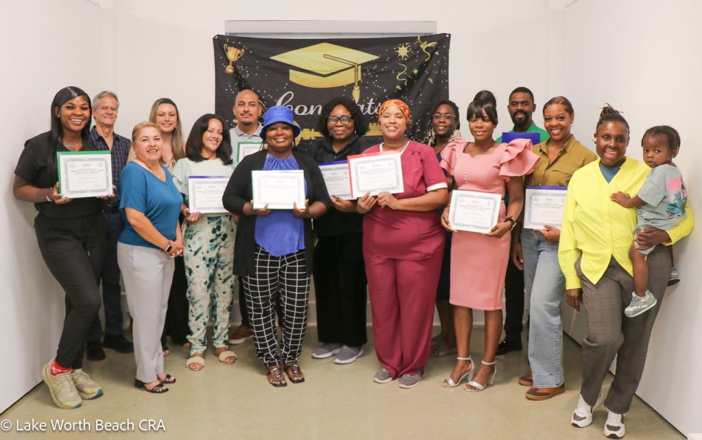 Community Partners of South Florida-Lake Worth Beach CRA Home Buyers Club Graduation A group of diverse graduates smiling and holding completion certificates in front of a celebratory graduation banner at a Community Partners of South Florida-Lake Worth Beach CRA Home Buyers Club event.