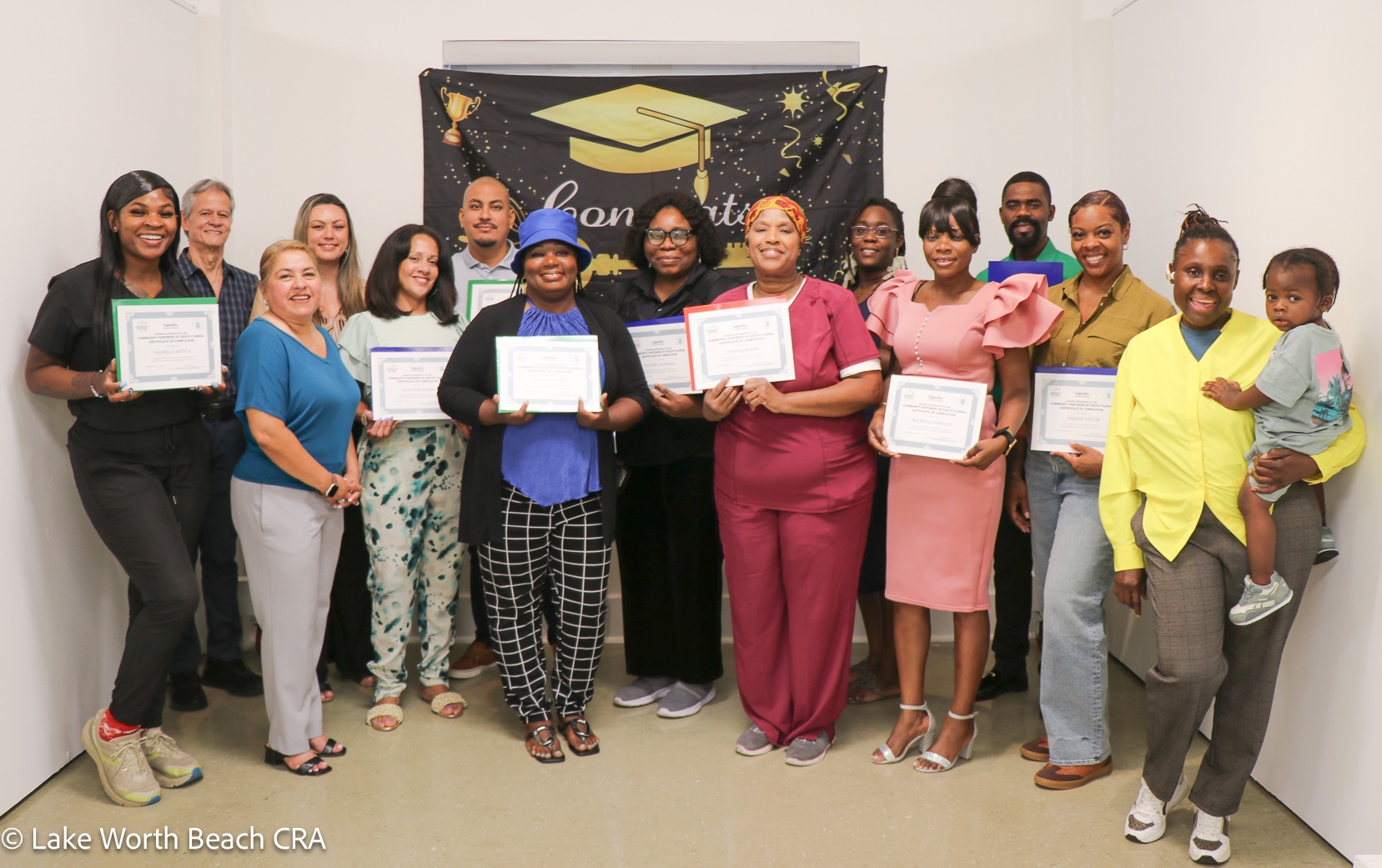 A group of diverse graduates smiling and holding completion certificates in front of a celebratory graduation banner at a Community Partners of South Florida-Lake Worth Beach CRA Home Buyers Club event.
