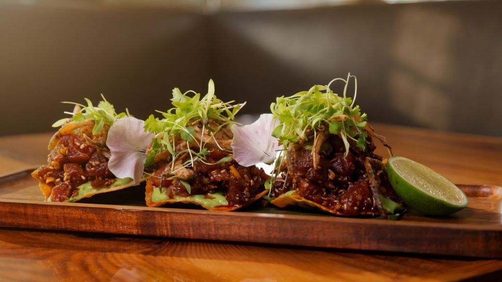 Three gourmet raw tuna tostadas garnished with microgreens and edible flowers served on a wooden board with a fresh lime wedge.