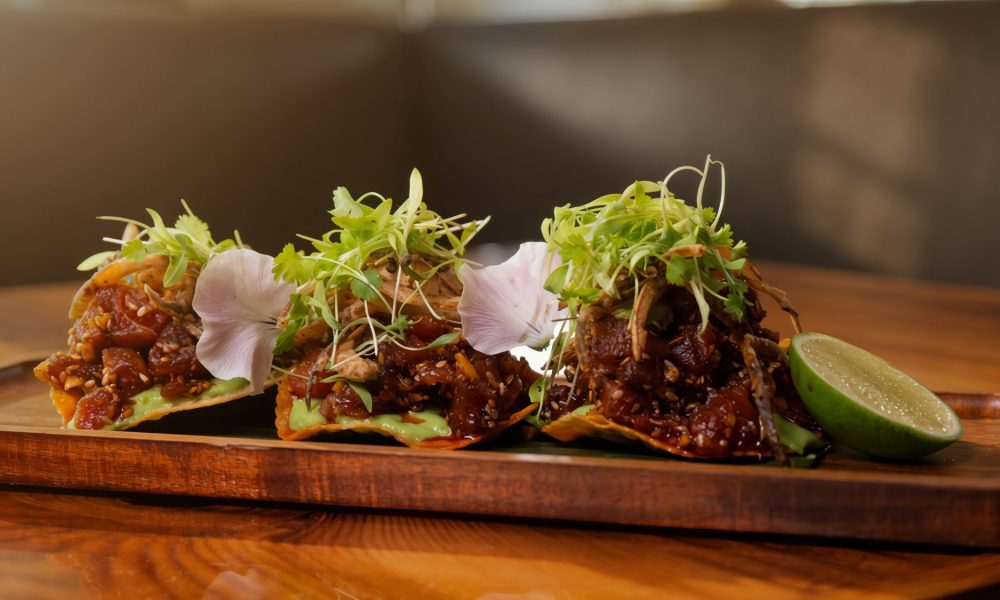 Three gourmet raw tuna tostadas garnished with microgreens and edible flowers served on a wooden board with a fresh lime wedge.