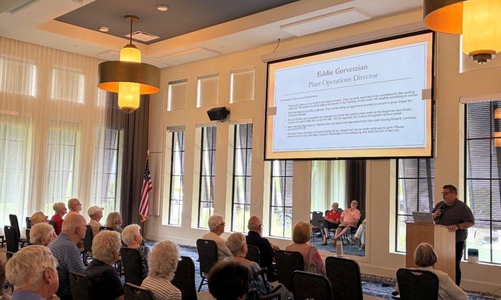 Wellington Bay Plant Operations Director presenting a hurricane readiness slide to a seated audience of senior residents inside a brightly lit luxury community clubhouse.