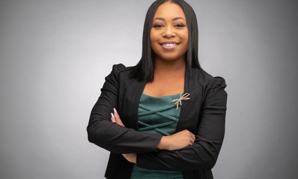 Headshot of Jade Lawson smiling with arms crossed, wearing a black blazer over a green dress with a dragonfly brooch.