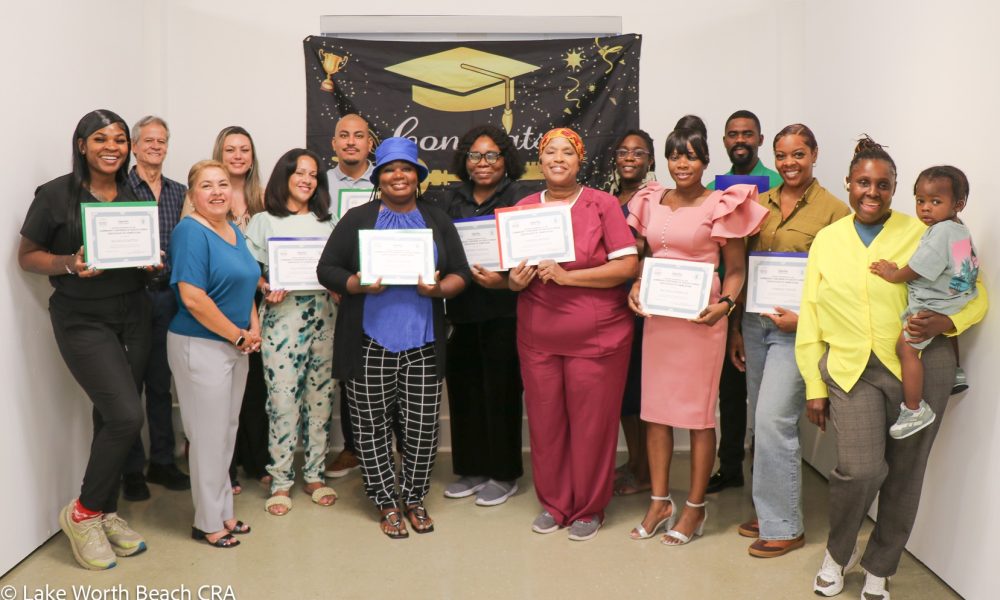 A group of diverse graduates smiling and holding completion certificates in front of a celebratory graduation banner at a Community Partners of South Florida-Lake Worth Beach CRA Home Buyers Club event.