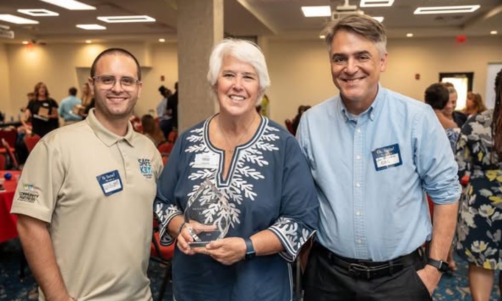 Three smiling individuals, including Kathy holding a crystal award, posing together at the FAU CARD Autism Leaders Brunch in Palm Beach County.